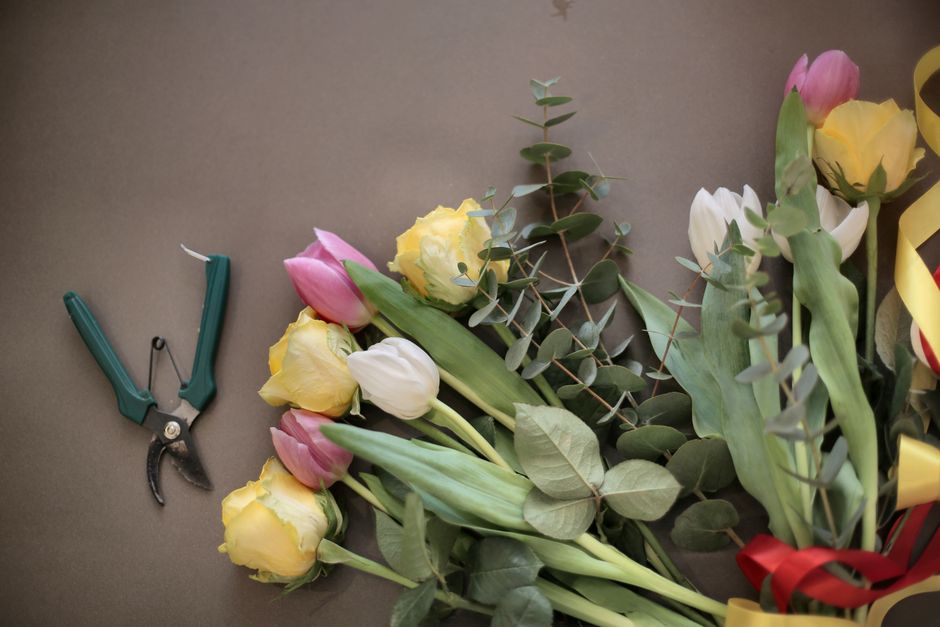 A floral arrangement featuring yellow, pink, and white tulips with lush green eucalyptus leaves laid out on a flat surface. A pair of green floral scissors is positioned nearby, indicating the process