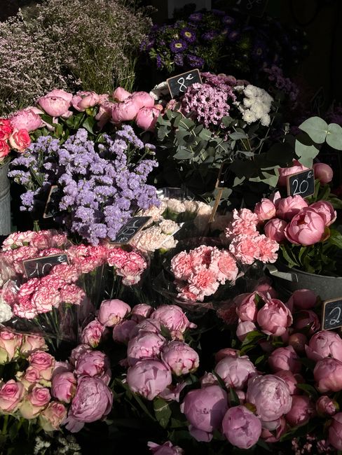 A vibrant display of fresh flowers arranged in bouquets and containers at a florist shop. The arrangement includes pink peonies with ruffled petals, purple statice with small delicate blossoms, white 