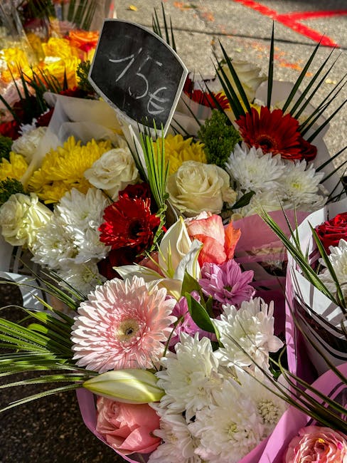 A vibrant bouquet of fresh flowers arranged in a pink wrapping, featuring large pink gerbera daisies, white roses, bright red gerbera daisies, and white chrysanthemums. The bouquet is displayed outdoo