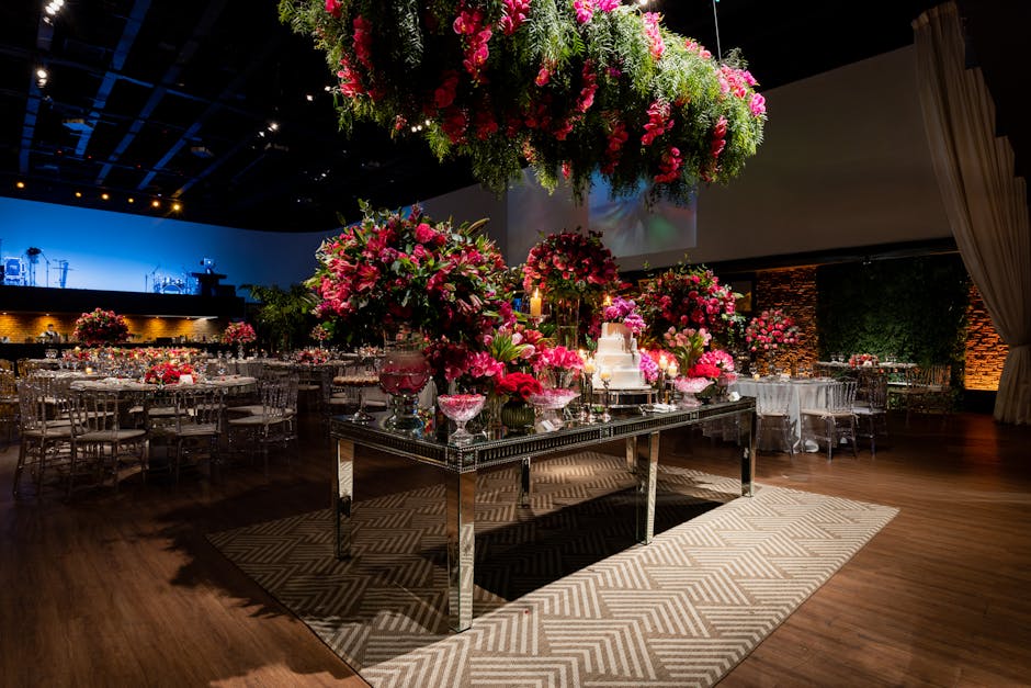 A floral arrangement featuring a large, elegant bouquet of pink roses, peonies, and lilies with green foliage, placed prominently on a reflective mirrored table. The arrangement is complemented by sma