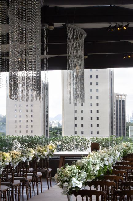 A floral arrangement featuring lush white hydrangeas, roses, and baby’s breath decorates a long banquet table in an elegant event space with large floor-to-ceiling windows revealing a cityscape of t
