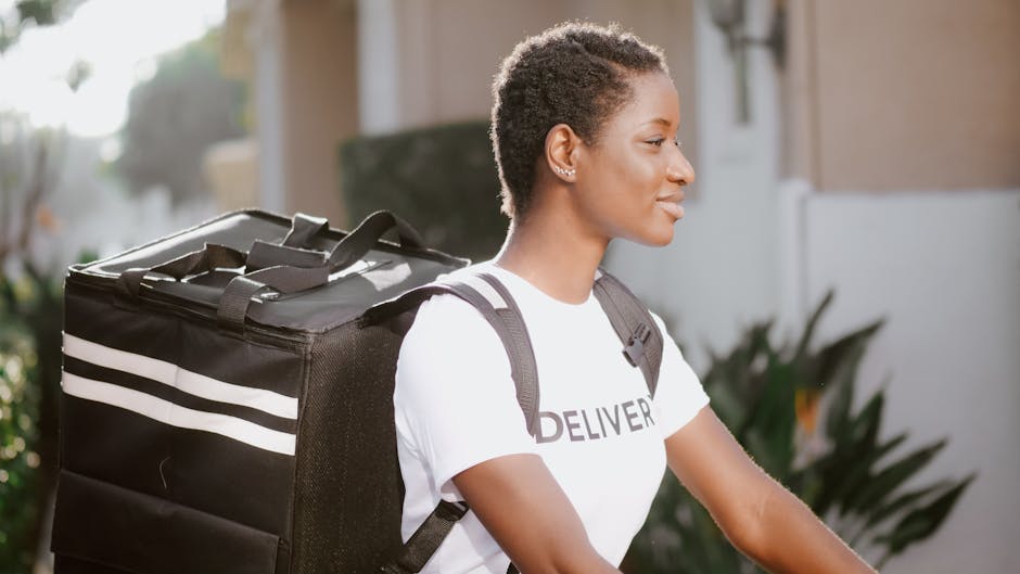 A young woman with short, curly hair and medium-dark skin is seen in profile, wearing a white t-shirt with the word 'DELIVER' printed on it, and a black backpack with white stripes. She is outdoors, s