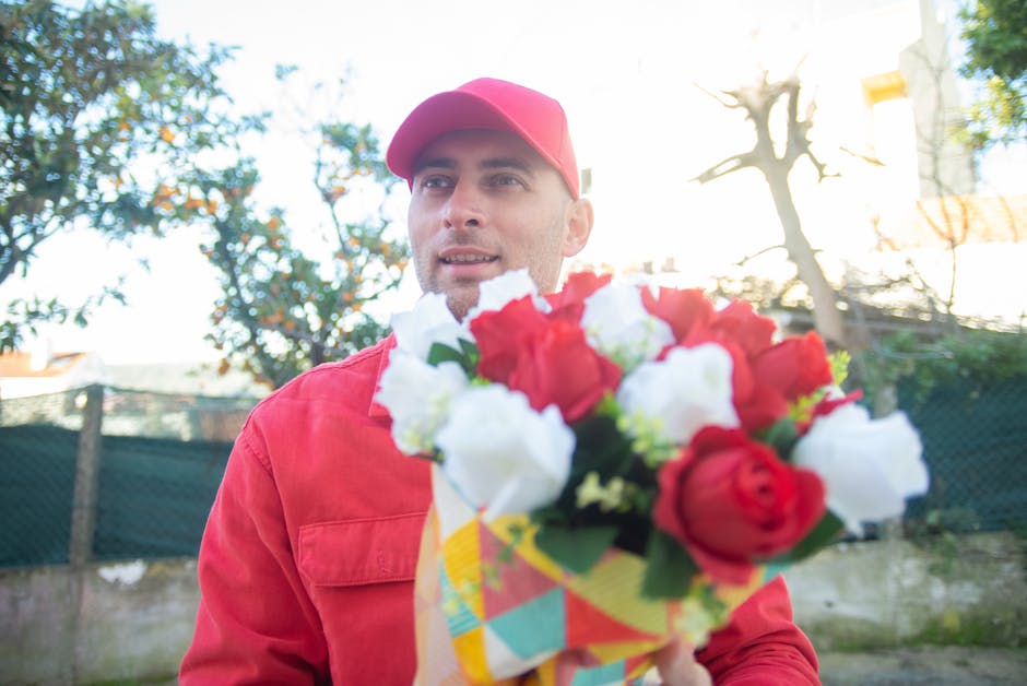 A male florist wearing a red cap and matching red jacket holding a colorful bouquet of fresh flowers, which includes red and white roses with green foliage, wrapped in patterned yellow, blue, and red 