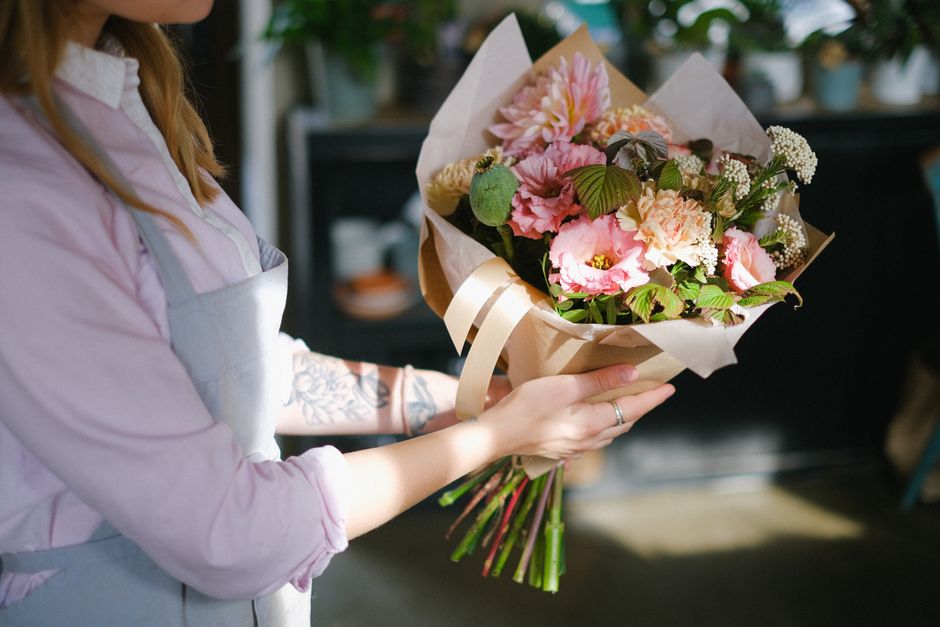 A person, wearing a light pink shirt and a grey apron, holds a beautifully arranged bouquet of fresh flowers. The bouquet includes large pink and cream carnations, delicate white baby's breath, and lu